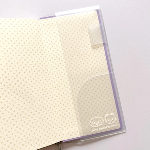 Op de afbeelding: Een transparante plastic notitieboekomslag met een paarse rand. De omslag heeft een zakje aan de rechterkant en een flap aan de linkerkant. De omslag heeft de tekst "Cute Stickers Only" gedrukt in de rechterbenedenhoek.