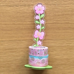 Op de afbeelding: Een set van drie roze en blauwe washi tapes met een bloemenpatroon. De tapes zijn gewikkeld rond een transparante plastic bloem met een roze strik. De bloem staat op een groene basis.