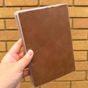 Op de afbeelding: Een bruin leren notitieboek met een doorzichtige plastic hoes. Het notitieboek wordt in een hand gehouden en de omslag heeft de tekst "SUNDAY MORNING" erop gedrukt. De achtergrond is een bakstenen muur.