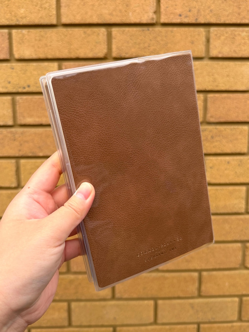 Op de afbeelding: Een bruin leren notitieboek met een doorzichtige plastic hoes. Het notitieboek wordt in een hand gehouden en de omslag heeft de tekst "SUNDAY MORNING" erop gedrukt. De achtergrond is een bakstenen muur.