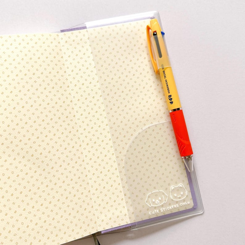 Op de afbeelding: Een doorzichtig plastic notitieboekje met een gele en oranje pen die eruit steekt. Het notitieboekje heeft een witte achtergrond met een patroon van kleine stippen. De pen heeft de tekst "HORIZON 1.0mm" en "B+B" erop staan. Het notitieboekje heeft de tekst "CUTE STICKERS ONLY" gedrukt in de rechterbenedenhoek.