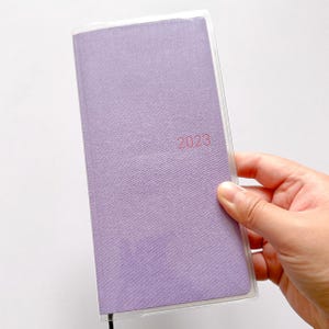 Op de afbeelding: Een paarse planner met een stoffen omslag en een transparante plastic hoes. Het jaar 2023 is in roze op de voorkant gedrukt.