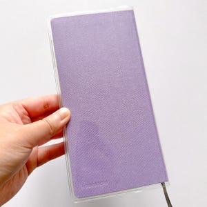 Op de afbeelding: Een lavendelkleurig notitieboekje met een transparante plastic hoes. Het notitieboekje heeft een textuur van stof en het woord "Hobonichi" staat op de voorkant gedrukt.