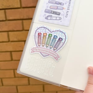 Op de afbeelding: Een doorzichtige planner met een vakje met stickers. E&eacute;n sticker toont kleurrijke potloden in een hartvorm met de tekst "My Favourite Rainbow". Een andere sticker toont verschillende penontwerpen.