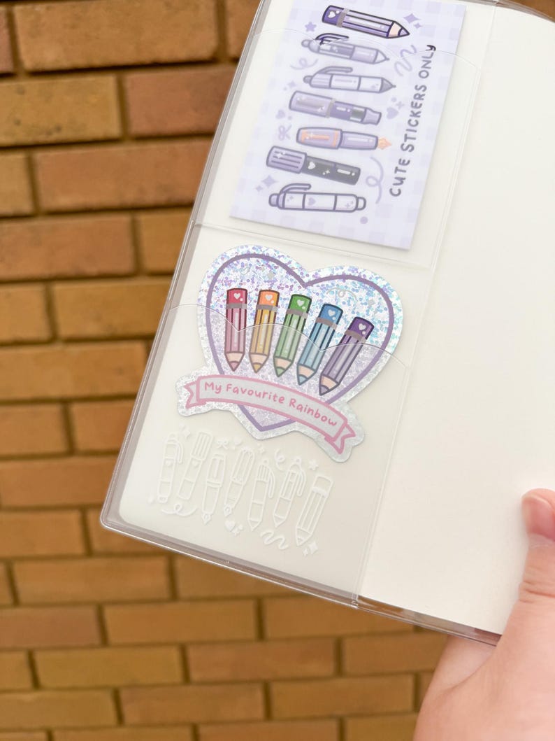Op de afbeelding: Een doorzichtige planner met een vakje met stickers. E&eacute;n sticker toont kleurrijke potloden in een hartvorm met de tekst "My Favourite Rainbow". Een andere sticker toont verschillende penontwerpen.