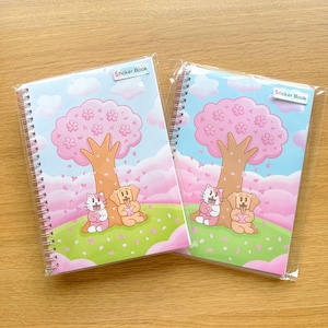 Op de afbeelding: Twee spiraalgebonden stickerboeken met een grillig ontwerp. Elk boek bevat een roze kersenbloesemboom, een kat en een hond op een groen veld onder een blauwe lucht. De tekst "Sticker Book" staat op elke omslag.