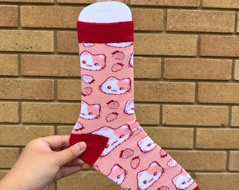 Op de afbeelding: Een roze sok met een herhaald patroon van witte kattengezichten en rode aardbeien. De sok heeft een rode manchet, hiel en teen. De sok wordt omhoog gehouden door een witte hanger.