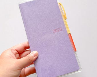 May include: A purple fabric-covered planner with a clear plastic cover. The planner has a black elastic band and the year 2023 printed on the front.