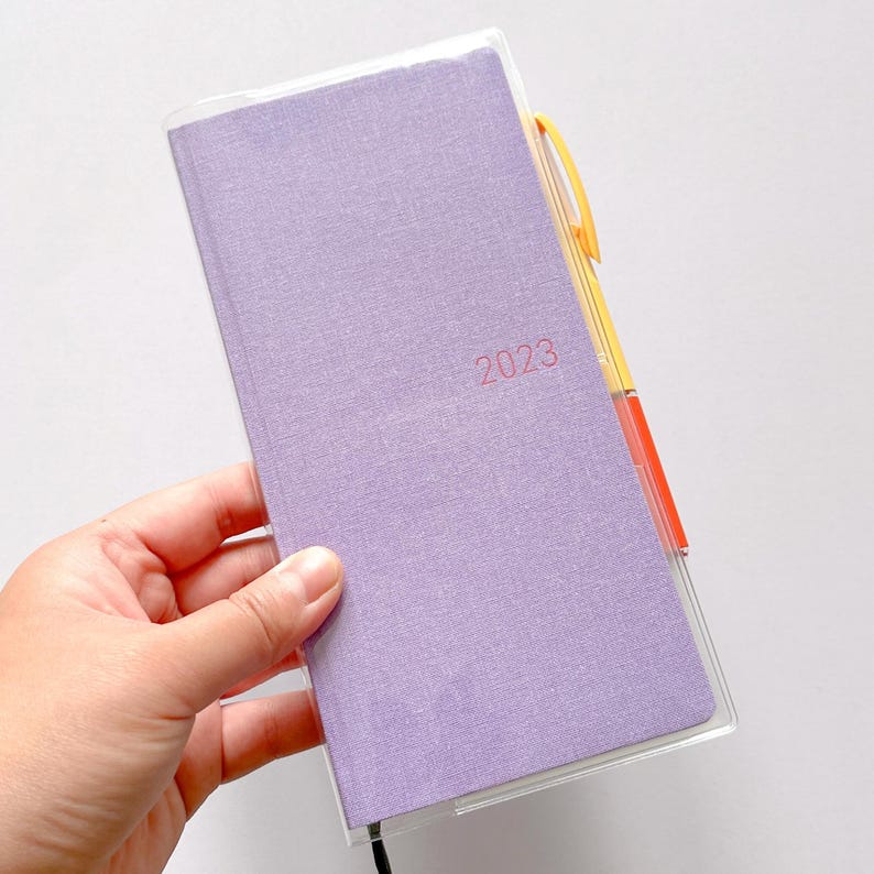Op de afbeelding: Een paarse planner met een stoffen omslag en een transparante plastic hoes. De planner heeft een zwart elastiek en het jaar 2023 staat op de voorkant gedrukt.