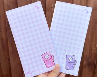 Op de afbeelding: Twee rechthoekige papieren notitieblokken met een lichtpaars en wit ruitpatroon. Elk notitieblok toont een kleine, cartoonachtige illustratie van een personage met een lachend gezicht, één in roze en één in paars, met kleine steraccenten.
