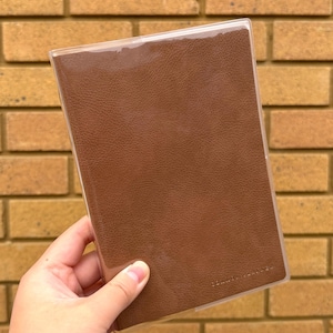 May include: A brown leather journal in a clear plastic cover. The journal is held in a person's hand. The background is a brick wall.