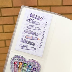 Op de afbeelding: Een doorzichtige plastic binder met twee sticker vellen. E&eacute;n vel bevat paarse pen illustraties en de tekst "Cute Stickers Only". De andere is een glitterhart met regenboog potlood illustraties en de tekst "My Favourite Rainbow."