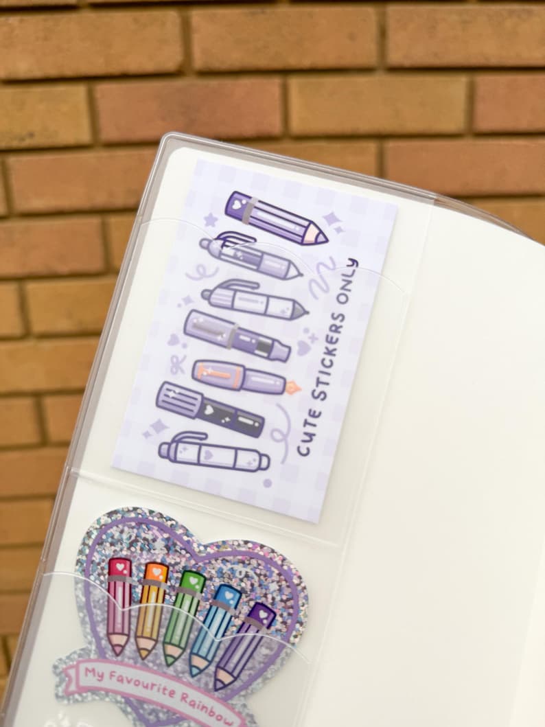 Op de afbeelding: Een doorzichtige plastic binder met twee sticker vellen. E&eacute;n vel bevat paarse pen illustraties en de tekst "Cute Stickers Only". De andere is een glitterhart met regenboog potlood illustraties en de tekst "My Favourite Rainbow."