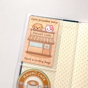 Op de afbeelding: Een transparante plastic zak met een sticker erin die "Cute Stickers Only" zegt. De sticker toont een cartooncaf&eacute; met een hond en een kat op het dak. De tekst "Kawaii Cafe" staat op de voorkant van het caf&eacute;. De sticker zegt ook "Have a lovely day!" Onder de zak zit een sticker met de tekst "Kawaii Cafe" met een koffiekopje en hartjes.