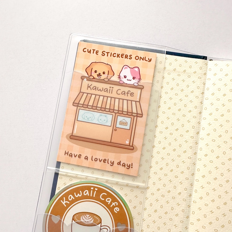 Op de afbeelding: Een transparante plastic zak met een sticker erin die "Cute Stickers Only" zegt. De sticker toont een cartooncaf&eacute; met een hond en een kat op het dak. De tekst "Kawaii Cafe" staat op de voorkant van het caf&eacute;. De sticker zegt ook "Have a lovely day!" Onder de zak zit een sticker met de tekst "Kawaii Cafe" met een koffiekopje en hartjes.
