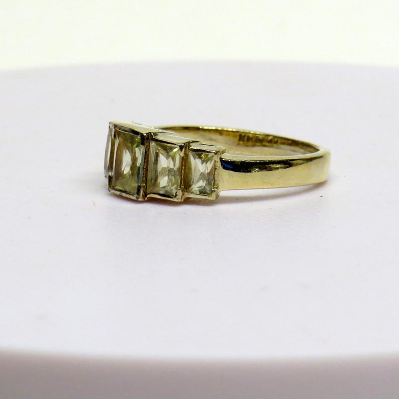 Op de afbeelding: Een gouden ring met drie rechthoekige, lichtgroene edelstenen. De band van de ring is van gepolijst goud en de edelstenen zijn in een rij geplaatst. De ring wordt gepresenteerd op een wit oppervlak, wat het ontwerp benadrukt.