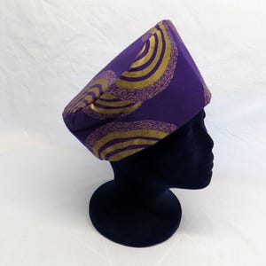 May include: A structured fabric hat in a deep purple hue, featuring a repeating pattern of concentric gold circles with a textured, stippled effect. The hat has a flat top and a wide, straight band that sits securely on a dark display head form.