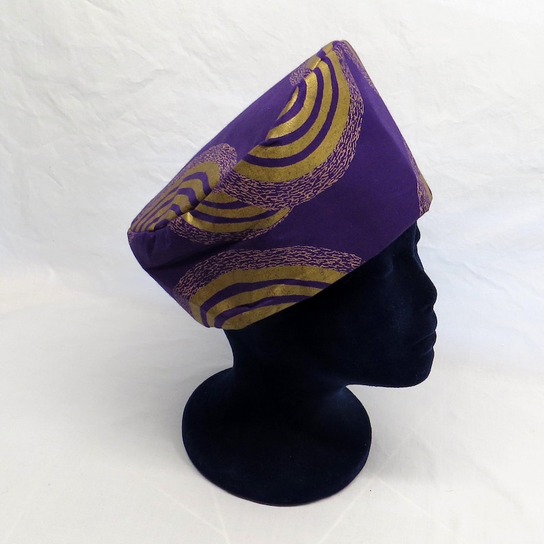 May include: A structured fabric hat in a deep purple hue, featuring a repeating pattern of concentric gold circles with a textured, stippled effect. The hat has a flat top and a wide, straight band that sits securely on a dark display head form.