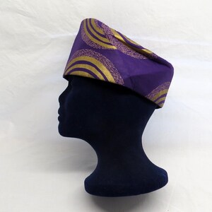 May include: A structured cap featuring a vibrant purple fabric base adorned with repeating gold circular patterns. The design consists of concentric gold arcs with textured detailing, creating a bold visual contrast against the deep purple background.