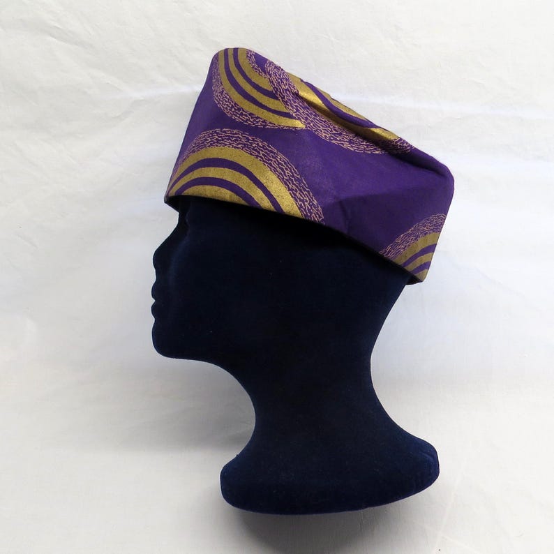May include: A structured cap featuring a vibrant purple fabric base adorned with repeating gold circular patterns. The design consists of concentric gold arcs with textured detailing, creating a bold visual contrast against the deep purple background.