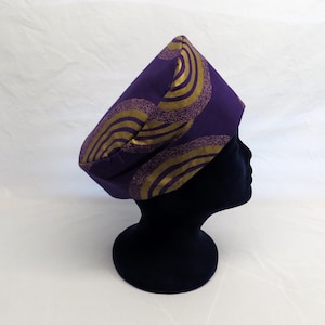 May include: A structured purple hat featuring a repeating pattern of gold-coloured concentric circles and textured, dark-toned curved lines. The headwear has a flat top and a wide, solid band that encircles the head, displaying a geometric design.