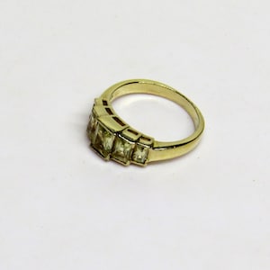 Op de afbeelding: Een goudkleurige ring met een reeks rechthoekige, lichtgroene edelstenen. De ring heeft een klassiek ontwerp met een gladde, gepolijste band en de edelstenen zijn in een lineair patroon geplaatst. De ring wordt op een wit oppervlak getoond.