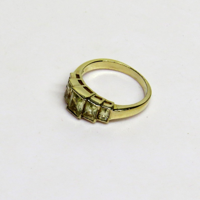 Op de afbeelding: Een goudkleurige ring met een reeks rechthoekige, lichtgroene edelstenen. De ring heeft een klassiek ontwerp met een gladde, gepolijste band en de edelstenen zijn in een lineair patroon geplaatst. De ring wordt op een wit oppervlak getoond.
