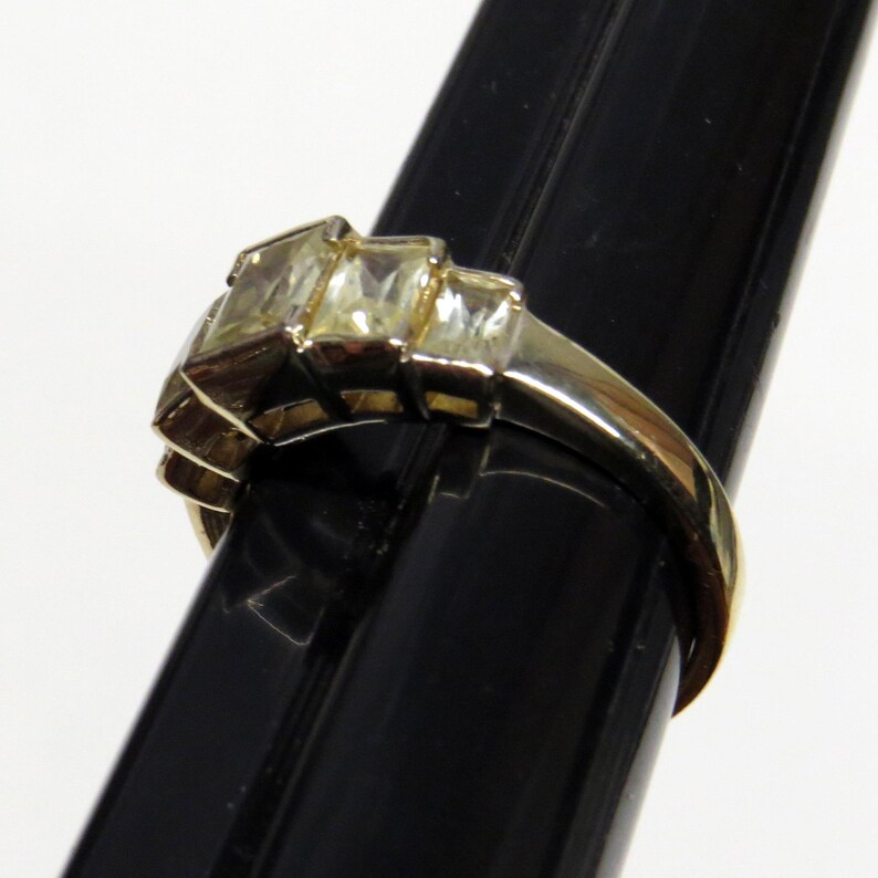 Op de afbeelding: Een goudkleurige ring met drie rechthoekige, heldere edelstenen in een trapsgewijs ontwerp. De ring wordt getoond op een zwarte standaard, die de gepolijste afwerking en ingewikkelde details benadrukt. De band heeft een glad, afgerond profiel.