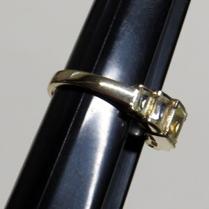 Op de afbeelding: Een gouden ring met drie vierkant geslepen, lichtgele edelstenen. De ring wordt getoond op een zwarte standaard, die het ontwerp en de glans van de stenen benadrukt. De ring heeft een gladde, gepolijste band.