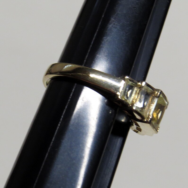 Op de afbeelding: Een gouden ring met drie vierkant geslepen, lichtgele edelstenen. De ring wordt getoond op een zwarte standaard, die het ontwerp en de glans van de stenen benadrukt. De ring heeft een gladde, gepolijste band.