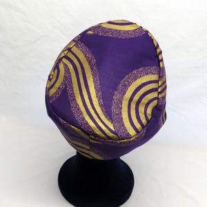 May include: A rounded cap featuring a deep purple base colour with a prominent gold metallic pattern consisting of swirling, concentric circular lines and textured bands that wrap around the entire surface of the headwear piece.