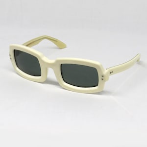 May include: Off-white rectangular sunglasses with dark lenses. The frames are thick and have a slight curve, with a lighter-colored arm. The sunglasses have a retro, vintage aesthetic.