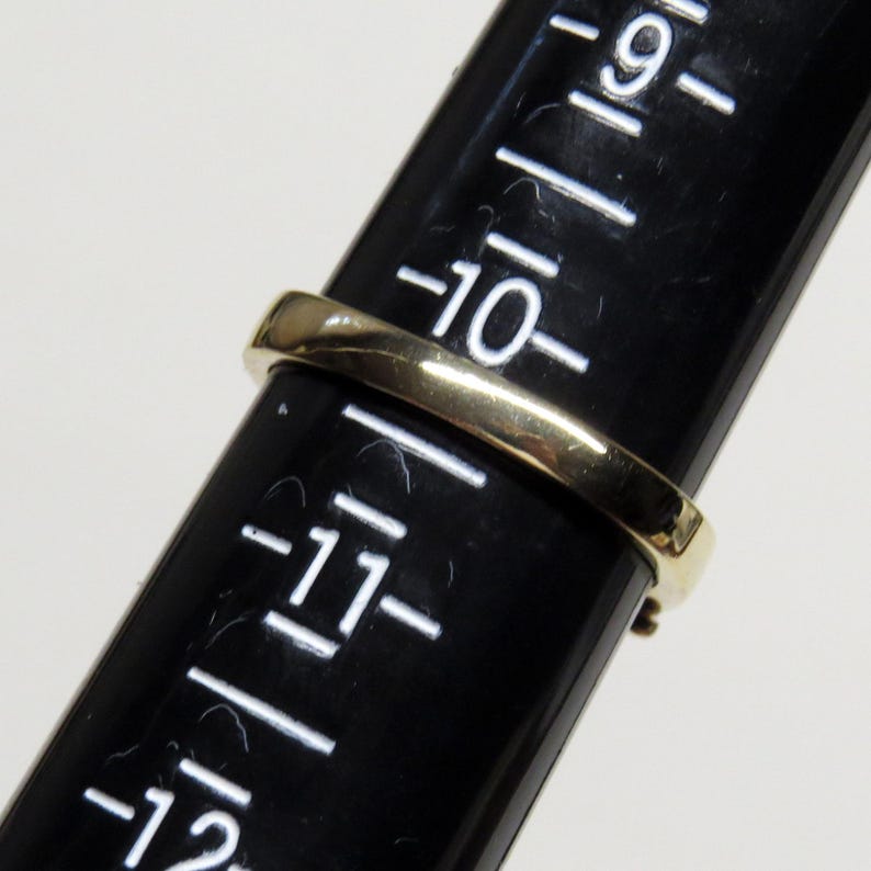 Op de afbeelding: Een gouden ring wordt op een zwarte ringmaat geplaatst. De ring heeft een eenvoudig bandontwerp. De ringmaat heeft witte markeringen en cijfers, waaronder het nummer 10. De ring wordt op maat gemeten.