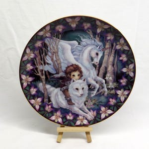 May include: Decorative plate featuring a fantasy scene. A child rides a white wolf, with a unicorn in the background. The plate has a gold rim and a floral border with purple and pink flowers. The background is a forest scene.