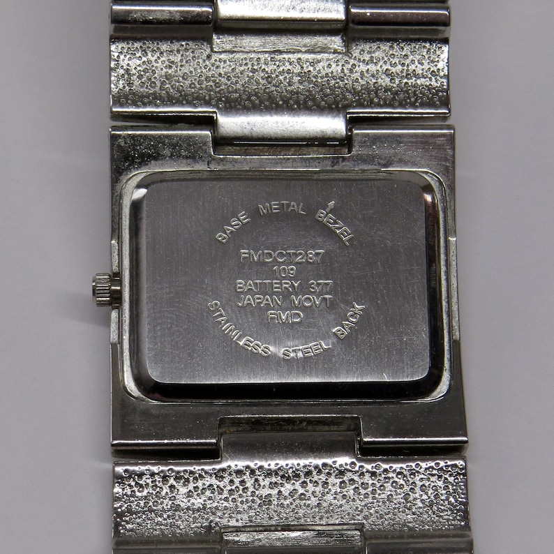 Może przedstawiać: Zbliżenie na tył stalowego zegarka. Na tylnej części zegarka widnieje napis "BASE METAL BEZEL", "FMDCT287 109 BATTERY 377 JAPAN MOVT RMD STAINLESS STEEL BACK". Pasek zegarka jest teksturowany.