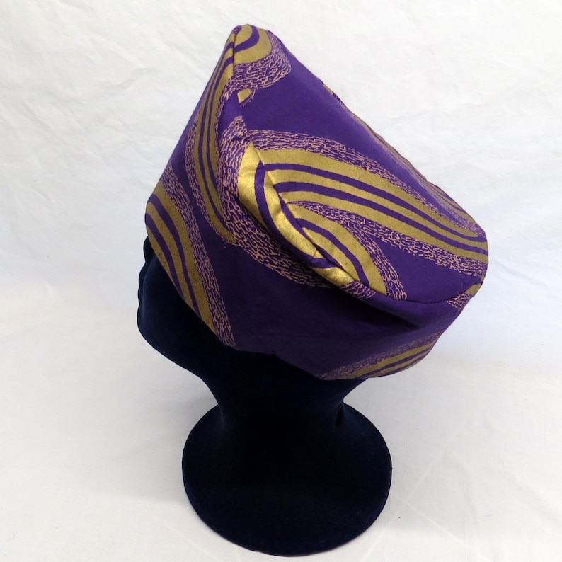 May include: A structured cap featuring a deep purple base colour with a prominent gold and textured light purple swirling pattern. The hat has a distinct pointed crown and a rounded silhouette, displaying a bold contrast between the metallic gold lines and the dark purple fabric throughout the design.
