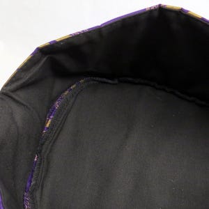May include: A circular fabric container with a black interior lining and a purple and gold patterned exterior border. The structure features a reinforced rim and a flat base, showcasing a contrast between the dark inner fabric and the colourful outer edge.