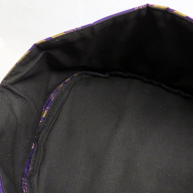 May include: A circular fabric container with a black interior lining and a purple and gold patterned exterior border. The structure features a reinforced rim and a flat base, showcasing a contrast between the dark inner fabric and the colourful outer edge.