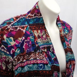May include: A long-sleeved jacket featuring a vibrant, multi-colored abstract pattern with shades of magenta, teal, deep purple, and black. The fabric has a distinct crinkled texture, a shawl collar, and an open front design without buttons or closures.