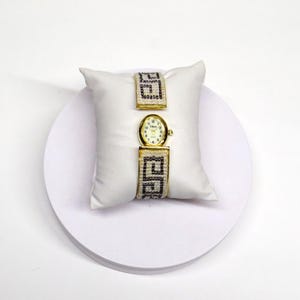 May include: A gold-toned wristwatch with a white face and black numerals, featuring a beaded band with a geometric design. The watch is displayed on a white satin pillow, showcasing its elegant design. The watch band is approximately 1 inch wide.