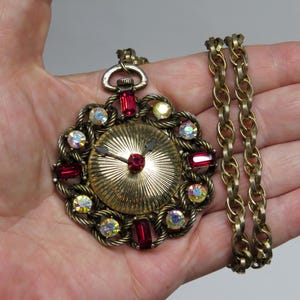 May include: Vintage gold-tone pendant necklace with a clock face design. The pendant features a gold-colored clock face with a red center stone and red and iridescent crystal accents. The necklace has a gold-colored chain.