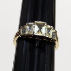 Op de afbeelding: Een goudkleurige ring met vijf rechthoekige, lichtgele edelstenen. De band van de ring is glad en gepolijst, met de edelstenen in een trapsgewijze opstelling. De ring wordt gepresenteerd tegen een zwarte achtergrond.