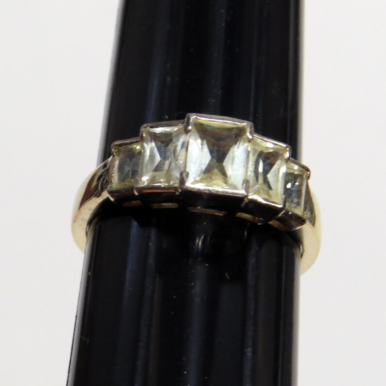 Op de afbeelding: Een goudkleurige ring met vijf rechthoekige, lichtgele edelstenen. De band van de ring is glad en gepolijst, met de edelstenen in een trapsgewijze opstelling. De ring wordt gepresenteerd tegen een zwarte achtergrond.