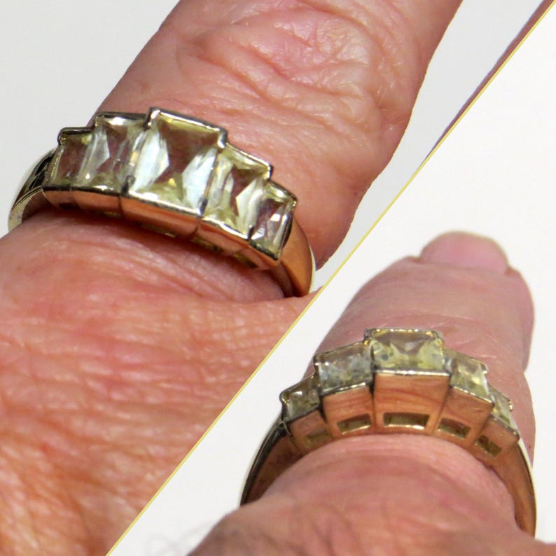 Op de afbeelding: Een goudkleurige ring met vijf rechthoekige, heldere edelstenen. De ring heeft een gepolijste gouden band. De edelstenen zijn in een trapsgewijs patroon gerangschikt, met de grootste steen in het midden.