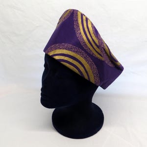 May include: A structured purple hat featuring a repeating pattern of gold concentric circles and textured gold lines. The hat is displayed on a dark, featureless mannequin head, showcasing its pointed, angular shape and vibrant colour contrast.