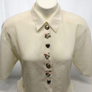 May include: Cream-colored short-sleeve blouse with decorative buttons. The buttons include a pearl, heart, and floral designs. The blouse is displayed on a mannequin.