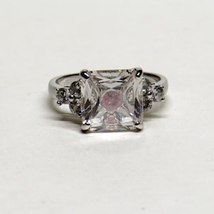 May include: A silver ring with a large, square, clear gemstone as the centerpiece. Smaller, round, clear gemstones are set on either side of the main stone. The ring is set against a white background.