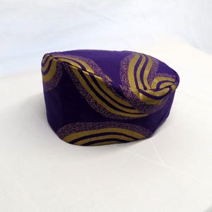 May include: A structured, round-top hat in a deep purple fabric featuring a repeating pattern of curved, golden-yellow lines with a textured, stippled appearance. The hat has a flat top and a short, vertical side band, showcasing a bold colour contrast.