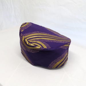 May include: A structured, brimless cap in a deep purple fabric featuring a repeating pattern of curved, metallic gold lines. The design consists of sweeping, wave-like gold bands that contrast against the solid purple background of the headwear.