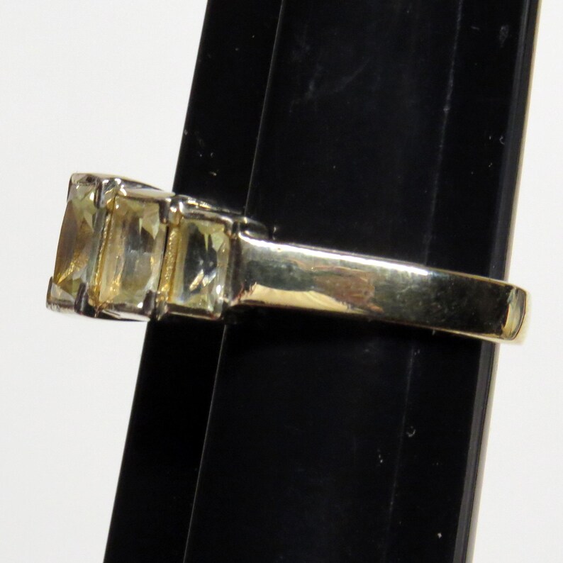 Op de afbeelding: Een goudkleurige ring met drie rechthoekige, lichtgele edelstenen. De ring heeft een gladde, gepolijste band, met de edelstenen in een rij geplaatst. De ring wordt gepresenteerd tegen een zwarte achtergrond.